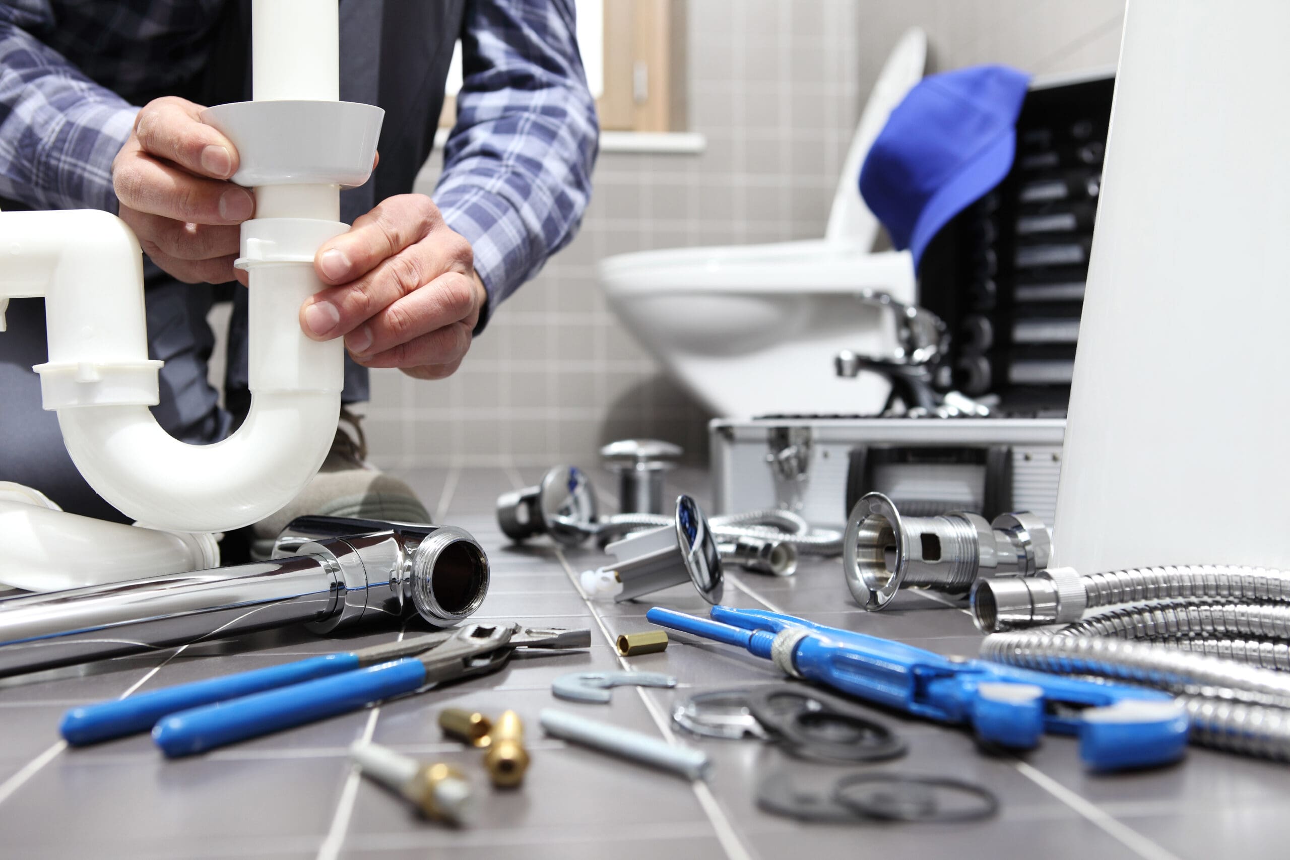 A plumber is fixing a pipe under a sink. Various tools and plumbing supplies are scattered on the tiled bathroom floor, including wrenches, flexible hoses, and fittings. A tool box is open in the background near a toilet.