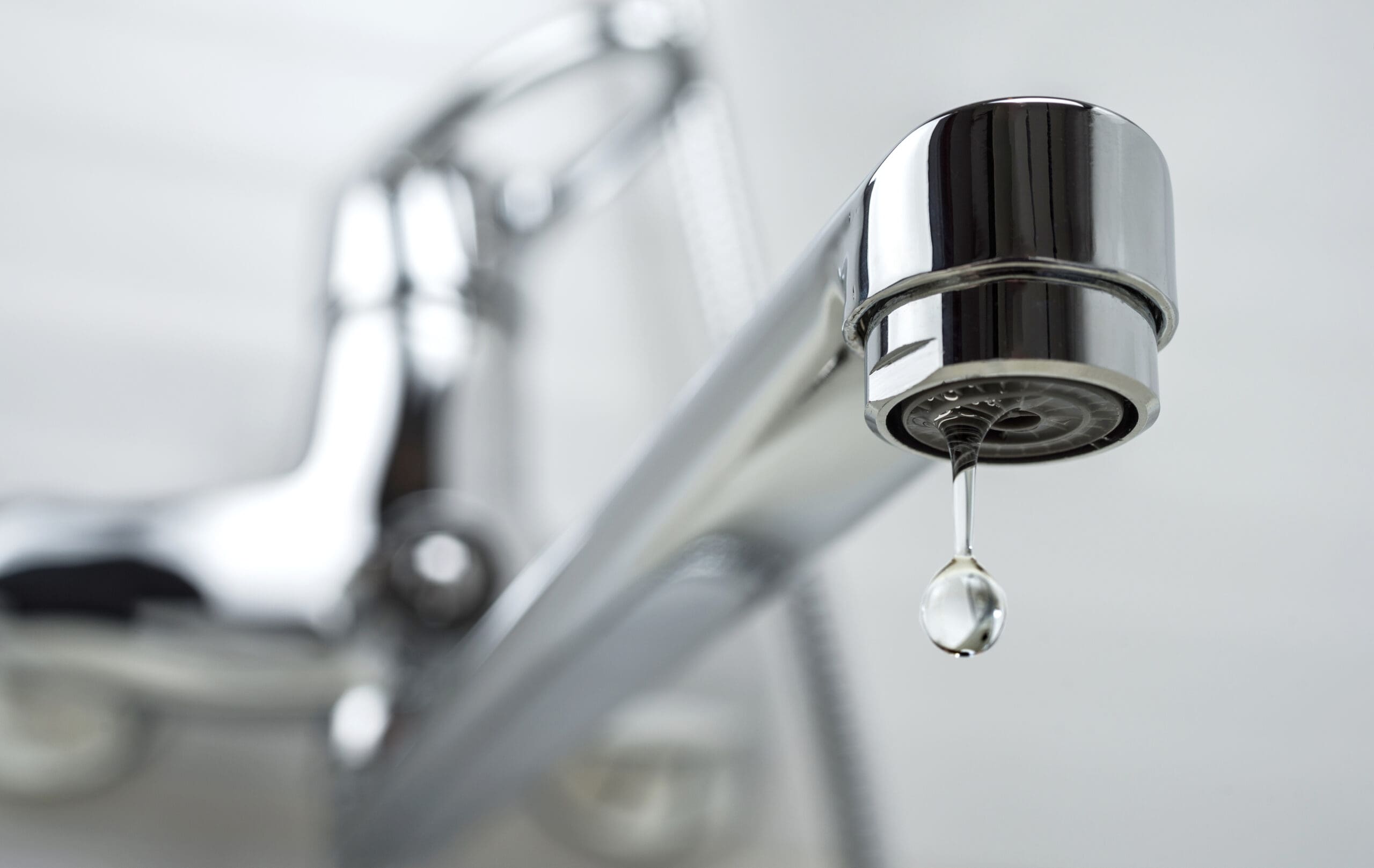 Close-up of a chrome faucet with a droplet of water about to fall from the spout. The background is blurry, highlighting the shiny surface and detail of the faucet and droplet.