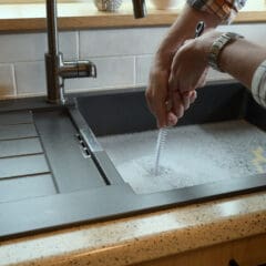 A person wearing a plaid shirt uses a brush to clean a glass in a kitchen sink filled with soapy water. The sink is modern with a chrome faucet and a drainer on the left side.