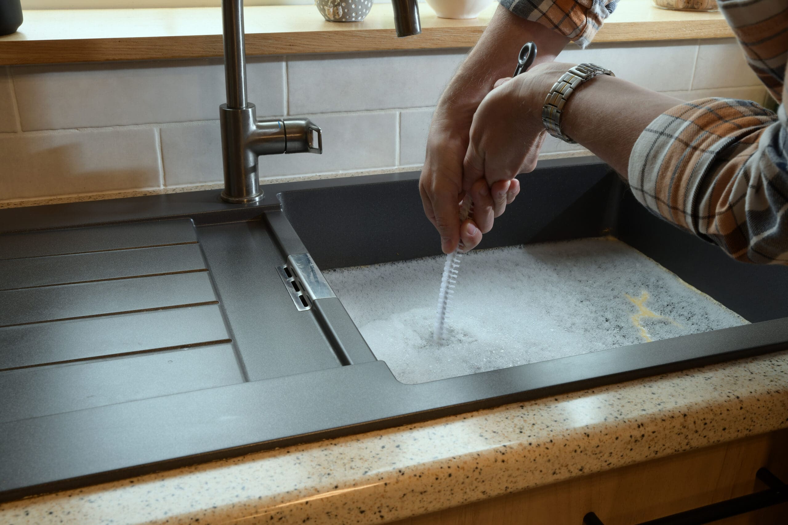 A person wearing a plaid shirt uses a brush to clean a glass in a kitchen sink filled with soapy water. The sink is modern with a chrome faucet and a drainer on the left side.