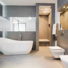 A modern bathroom featuring a sleek white freestanding bathtub, a shower with a glass partition, a wall-mounted toilet and bidet, and large mirrors reflecting natural light. The decor includes gray tiles and a minimalist design.