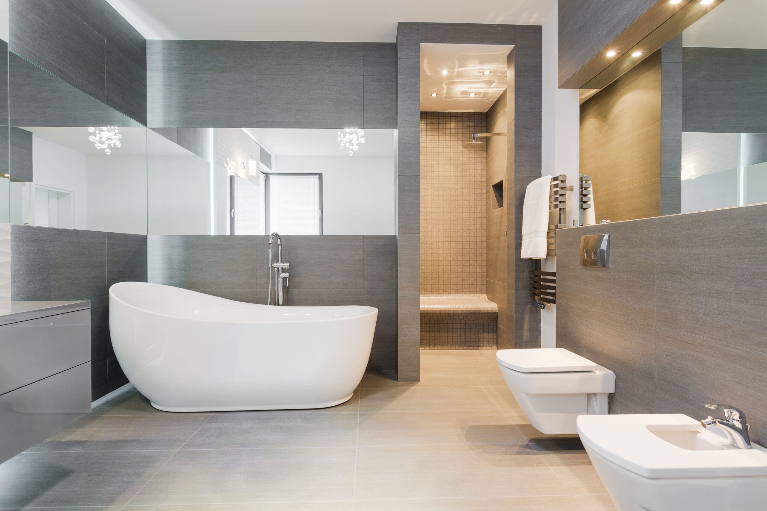 A modern bathroom featuring a sleek white freestanding bathtub, a shower with a glass partition, a wall-mounted toilet and bidet, and large mirrors reflecting natural light. The decor includes gray tiles and a minimalist design.