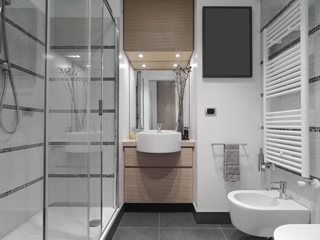 Modern bathroom with a glass shower on the left, a wooden vanity with a round sink and mirror in the center, and a bidet and towel rack on the right. The walls are tiled in white and gray, and the floor is dark tile.