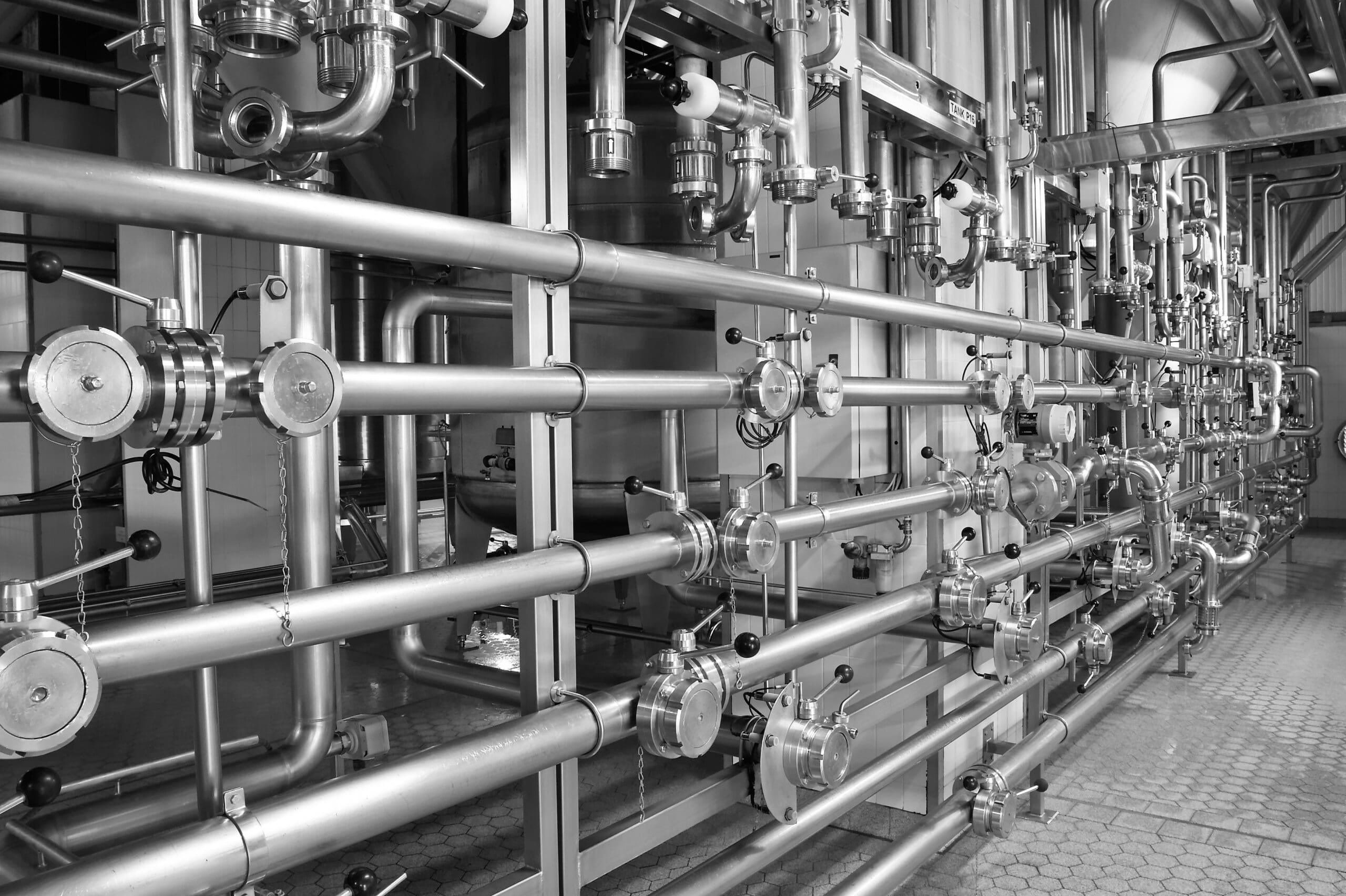 A complex network of stainless steel pipes and valves in an industrial facility. The pipes run horizontally and vertically, interconnected with circular valves, gauges, and other mechanical components on a textured floor. Image is in black and white.