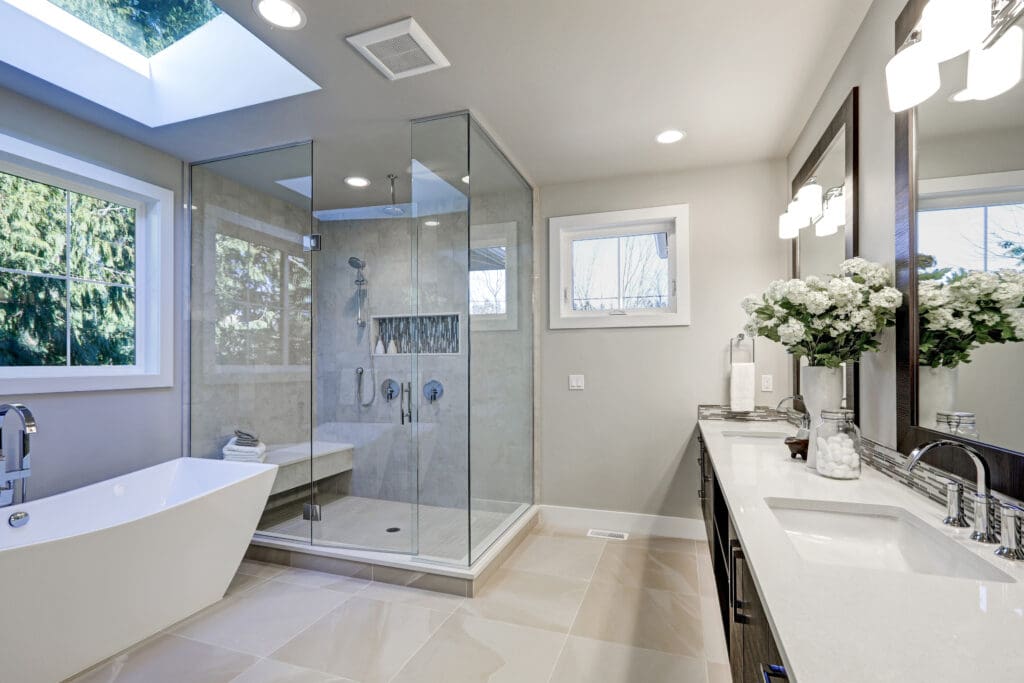 Modern bathroom with a freestanding bathtub, glass-enclosed shower, and dual-sink vanity. Large windows and a skylight illuminate the space. Decor includes a vase with flowers on the countertop. Neutral tones with polished surfaces.