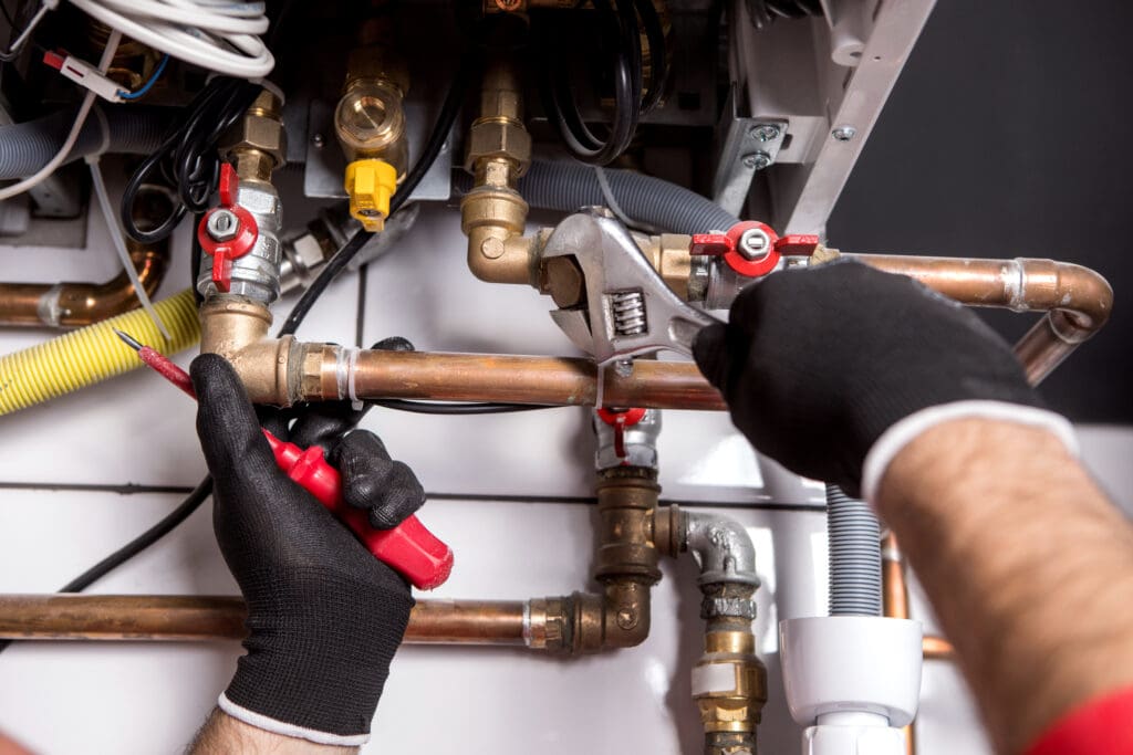 Close-up of a person's hands wearing black gloves using a wrench to tighten a pipe fitting. Another hand holds a red screwdriver near copper pipes and various valves, indicating a plumbing or maintenance task.