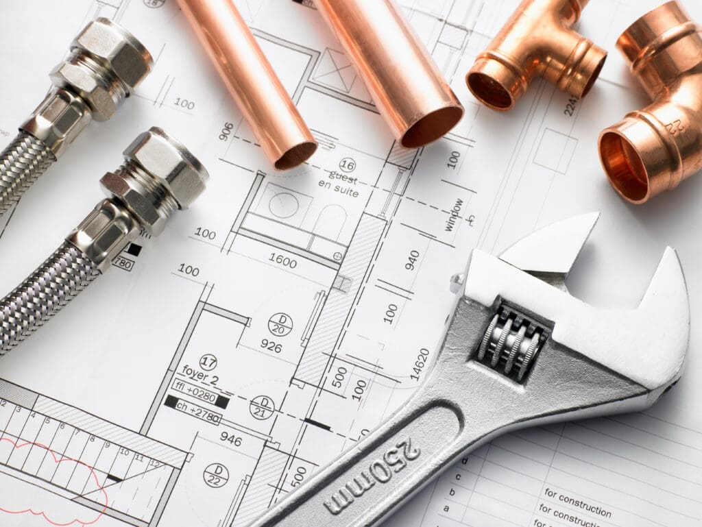 Plumbing tools and copper pipes are placed on an architectural blueprint. A large adjustable wrench lies in the foreground, with flexible hoses and pipe fittings scattered nearby, suggesting a focus on plumbing installation or repair.