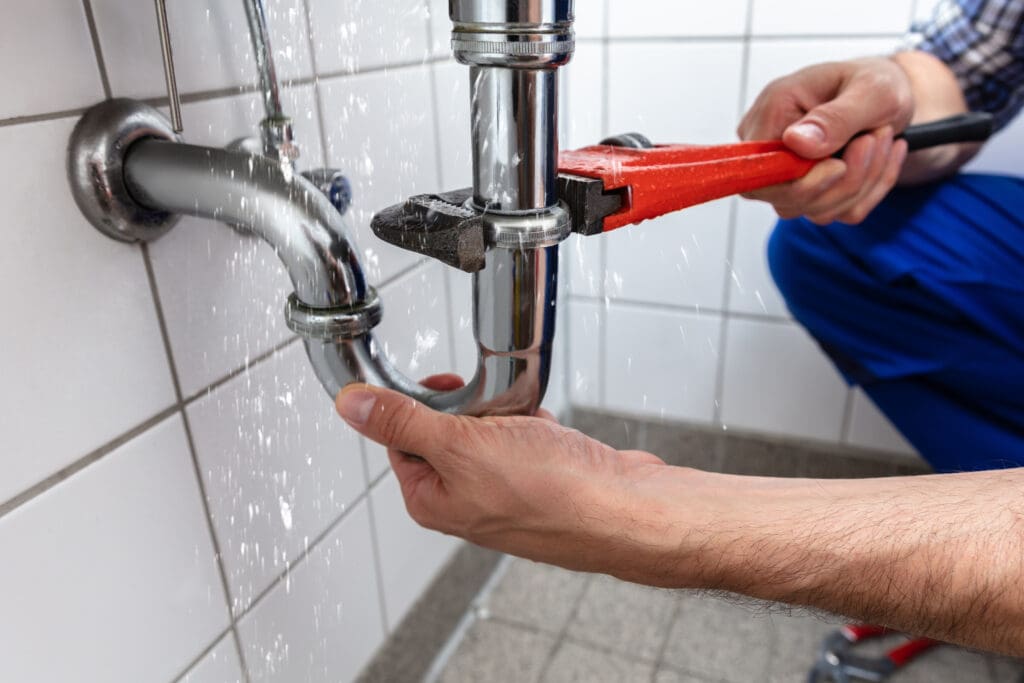 A person uses a red wrench to fix a leaking pipe under a sink. Water is spraying from the joint, wetting the tiled wall and floor. The background features white tiles, and the person wears blue pants.