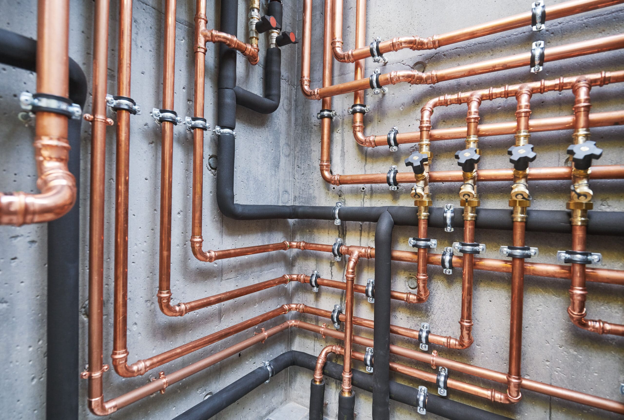 A complex network of copper and black insulated pipes are mounted on a concrete wall, featuring various joints and valves. The pipes intersect and run parallel at different angles, illustrating a detailed plumbing or heating system.