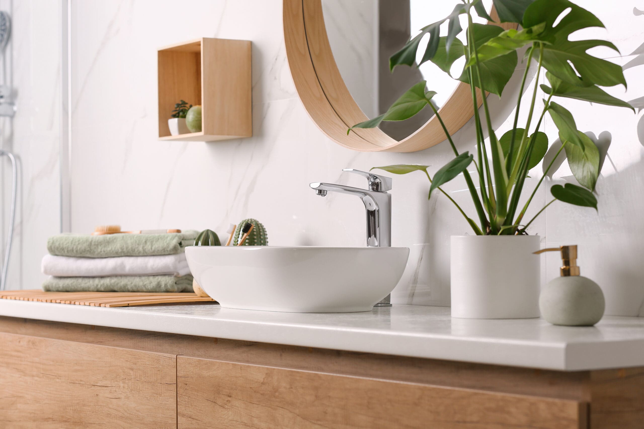 A modern bathroom features a white sink on a wooden countertop, a round mirror with a wooden frame, and a potted plant. Neatly folded towels and a soap dispenser are also present, offering a clean and stylish appearance.