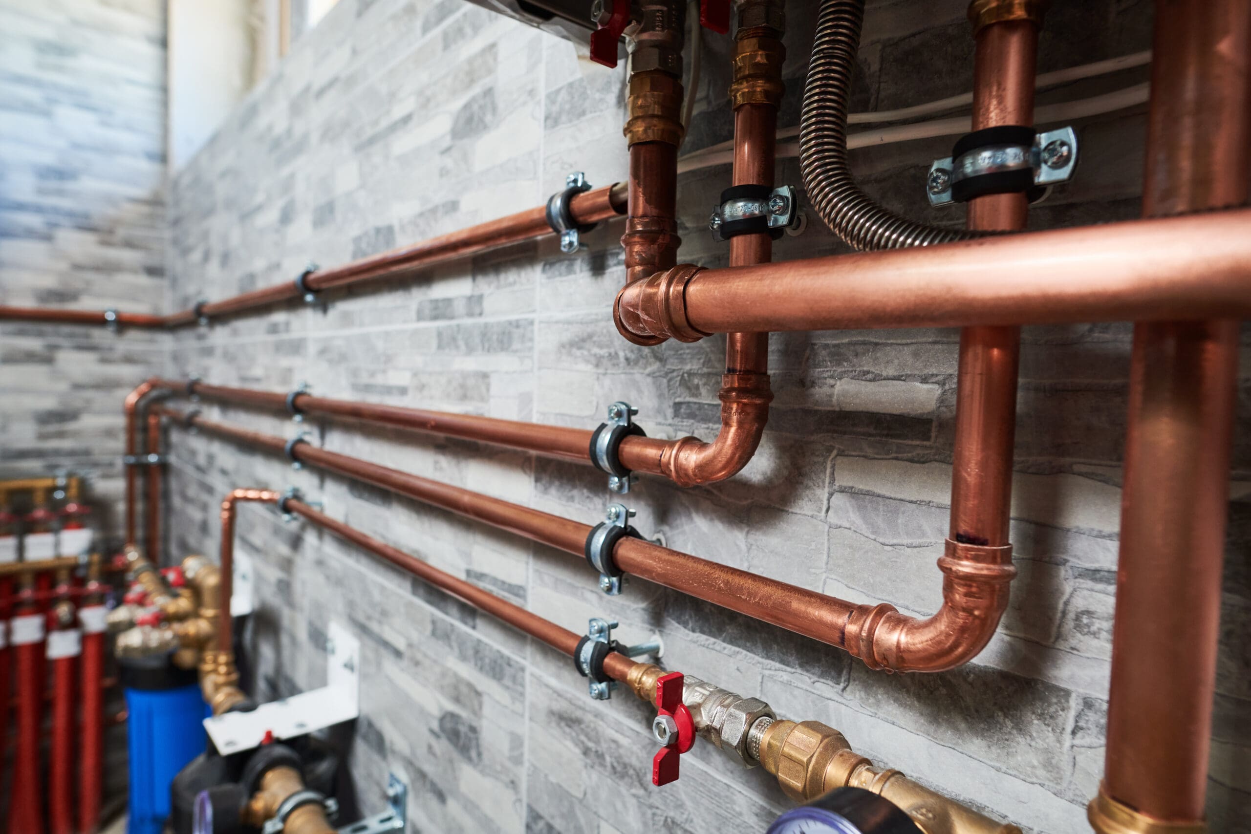 Copper pipes installed along a tiled wall, with various fittings, valves, and gauges. The pipes are secured with metal brackets, suggesting a plumbing or heating system in a utility area.