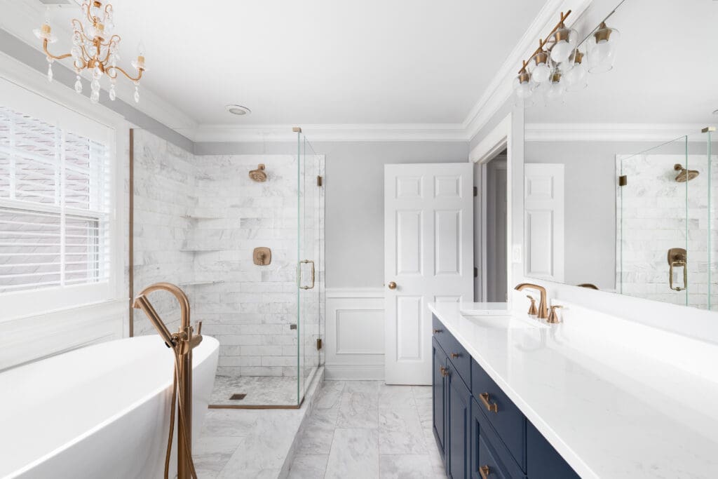 A modern bathroom with a freestanding bathtub, marble flooring, and a glass-enclosed shower. There are blue vanity cabinets with white countertops, and gold fixtures throughout. A chandelier hangs above, and a large window provides natural light.