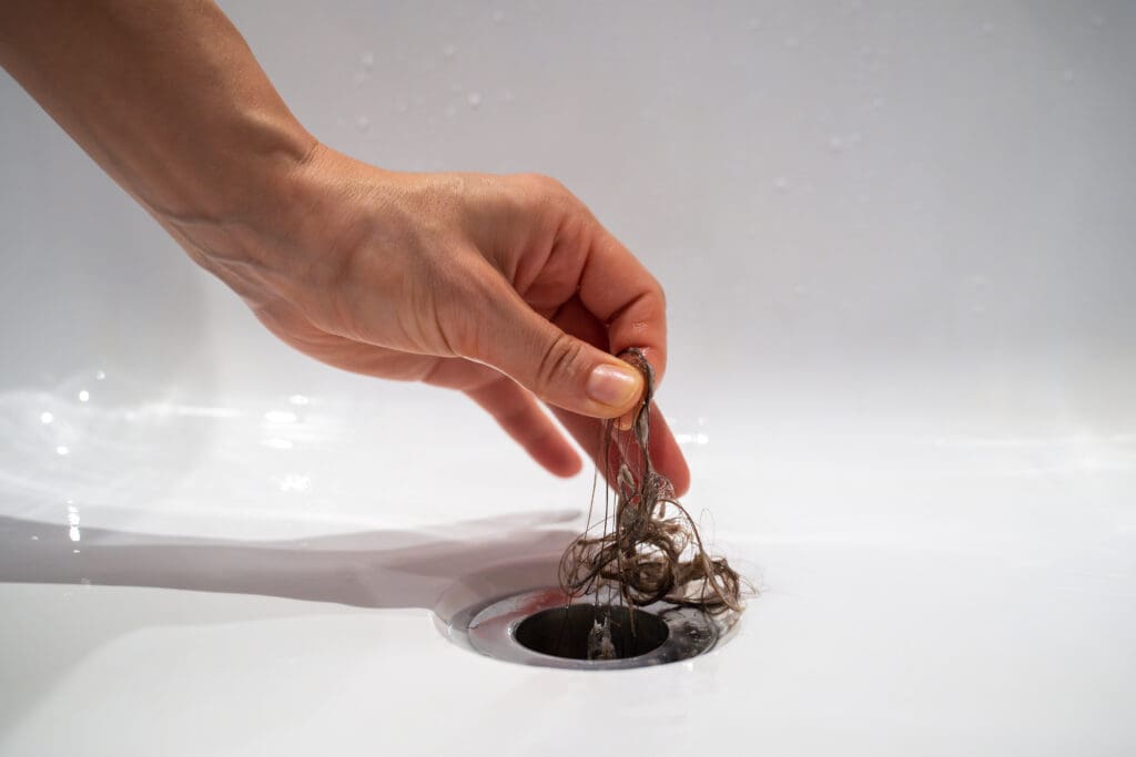 A hand is reaching into a bathroom sink drain, pulling out a clump of tangled hair. The white sink is free of water, and the focus is on the act of cleaning the drain.