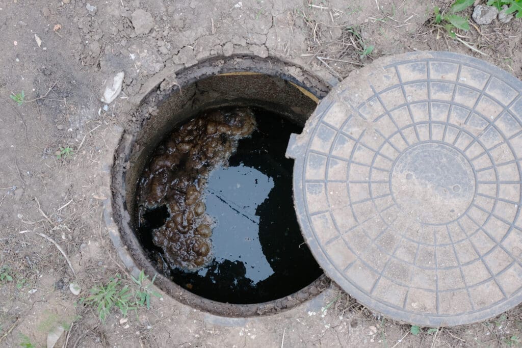 An open manhole with a partially moved round cover, exposing dirty water inside. The surrounding ground is dry and cracked, with patches of grass nearby.