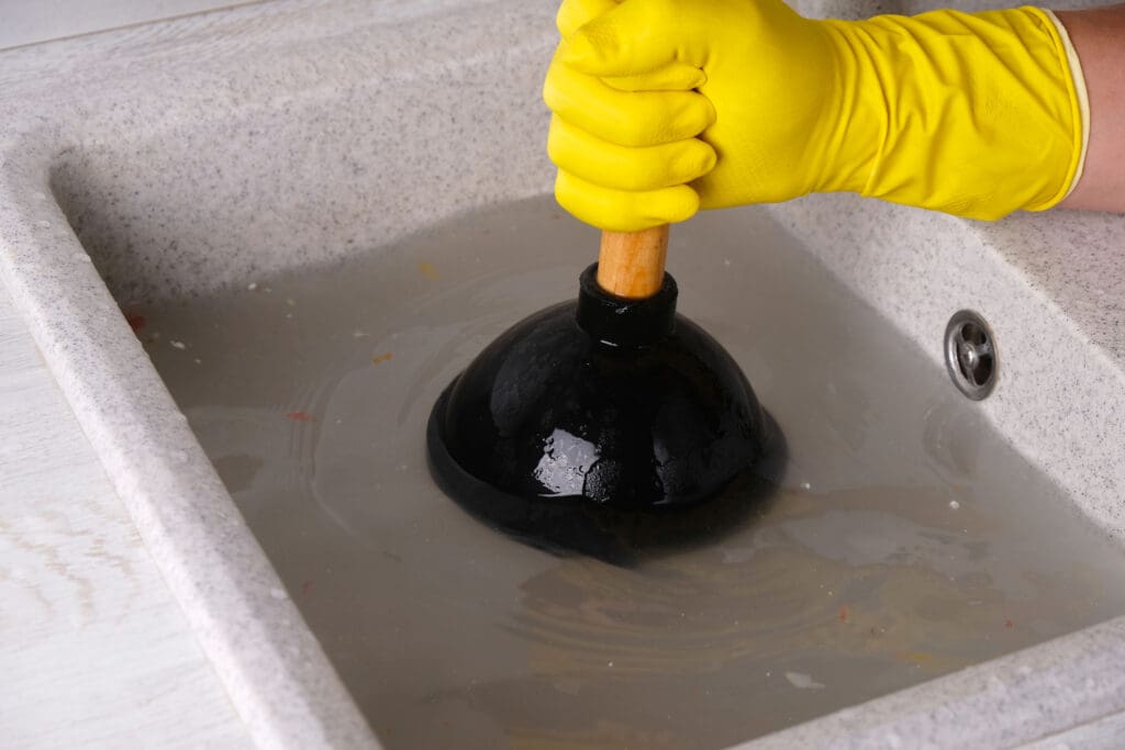 A person wearing yellow rubber gloves uses a plunger to unclog a sink filled with cloudy water.