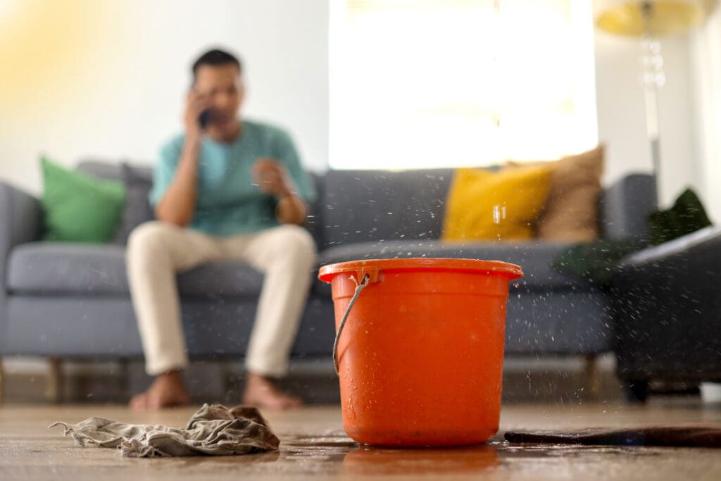 A blurry man sits on a couch talking on the phone, while an orange bucket sits on the floor catching water from a leak. A wet cloth lies nearby, and the scene suggests a leaking ceiling.