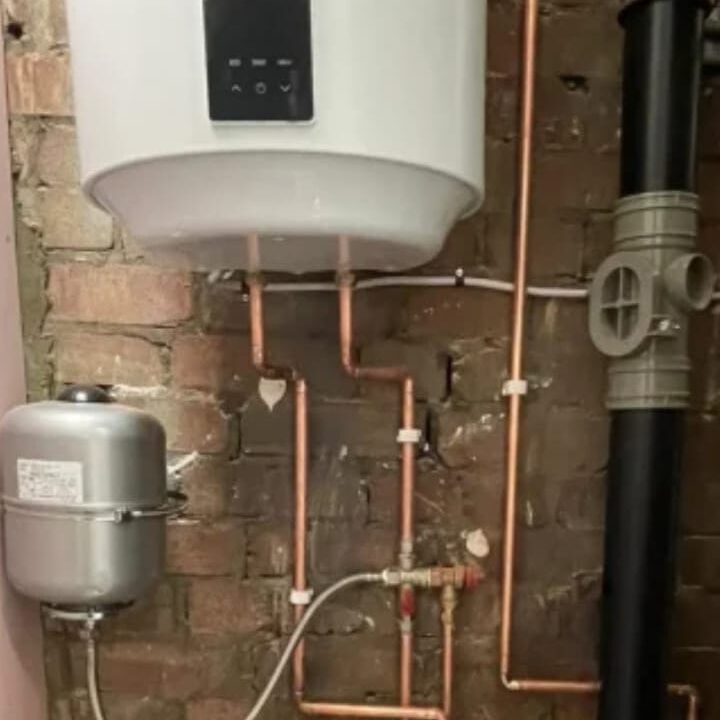 A water heater is mounted on a brick wall with copper piping connected. An expansion tank is positioned below, and a drainage pipe runs vertically nearby. The setup is part of a residential plumbing system.