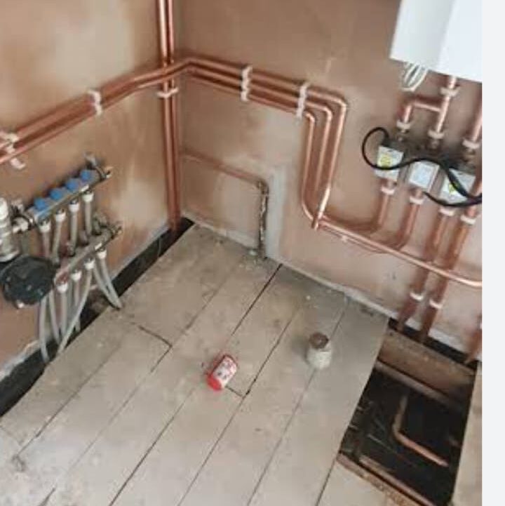 A partially completed plumbing installation with copper pipes on an unfinished wall. The floorboards are lifted, revealing some of the piping underneath. A small red can and a container are on the wooden floor.