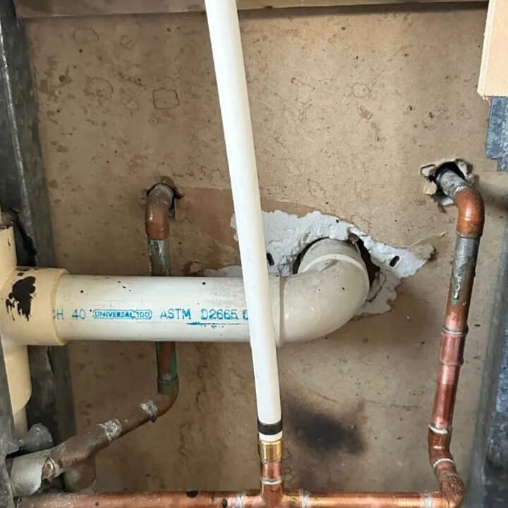 An exposed indoor plumbing system with PVC and copper pipes, including a section with visible wall damage where one pipe enters. The pipes are variously connected and show wear, against a wooden or particle board background.