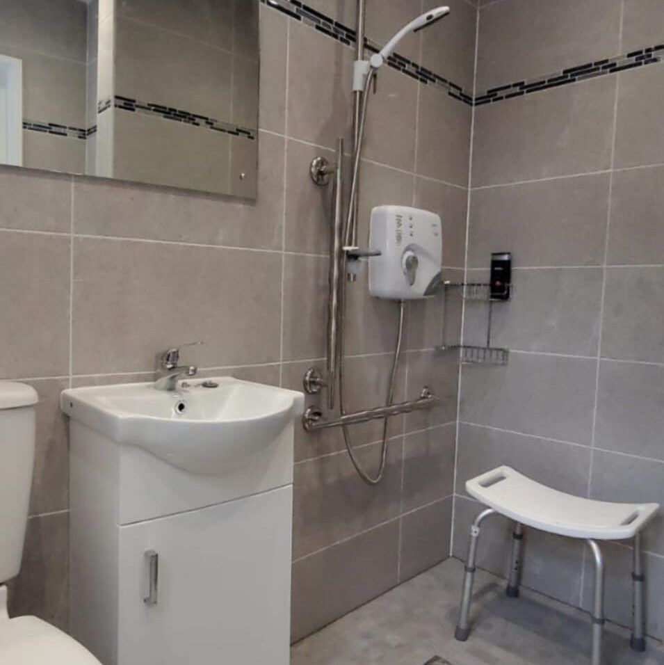 A modern bathroom with gray tiled walls features a walk-in shower equipped with a handheld showerhead and grab bars. A white sink with a cabinet is next to a toilet. A shower stool and wall-mounted soap dispenser are also present.