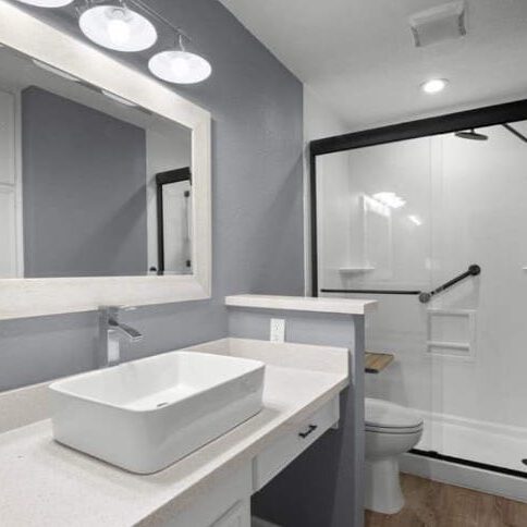 A modern bathroom featuring a large square sink on a white countertop, a frameless mirror, and a walk-in shower with a glass door. The walls are painted gray, and recessed lighting illuminates the space.