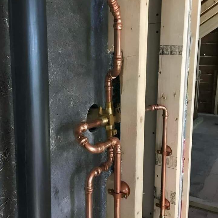 Copper plumbing pipes are installed inside an unfinished wall, showing connections and bends. They are set against wooden beams and a black surface, indicating the plumbing layout in early-stage construction.