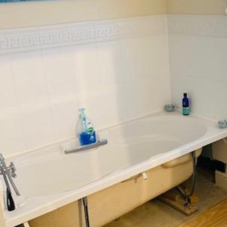 A white bathtub without a panel on one side, exposing the interior plumbing. Two bottles, one blue and one dark blue, are placed at opposite ends of the tub. The surrounding walls are white tiled.