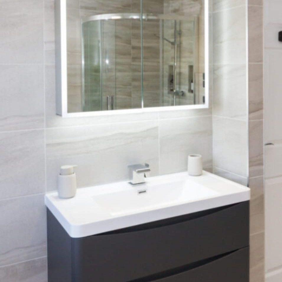 Modern bathroom featuring a dark-colored vanity with a white countertop and a rectangular sink. A backlit mirror hangs above the sink, reflecting a glass shower enclosure. The walls are tiled in light gray.