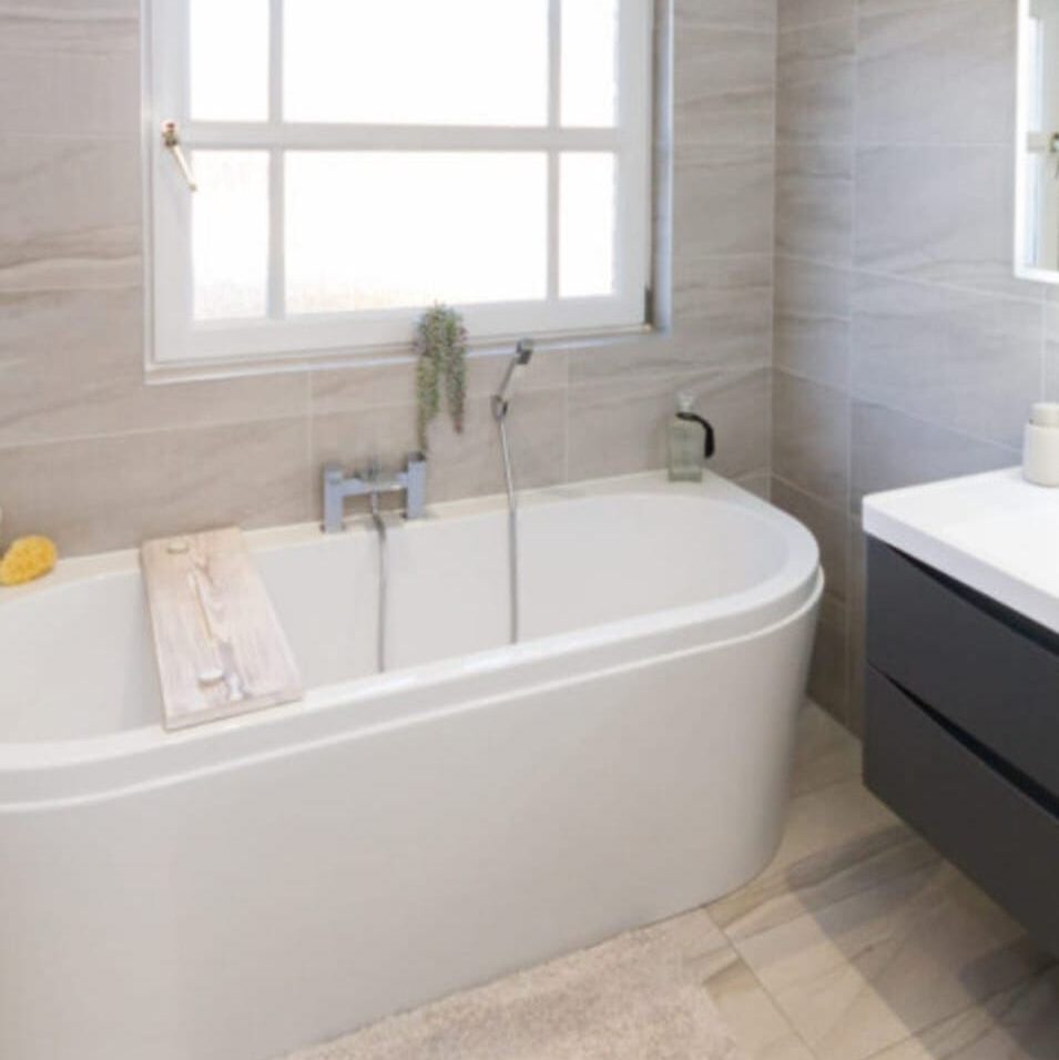 Modern bathroom with a white freestanding bathtub under a window. The tub has a wooden tray and a small plant on the edge. Gray tiled walls and flooring complement the dark vanity with a white countertop on the right.