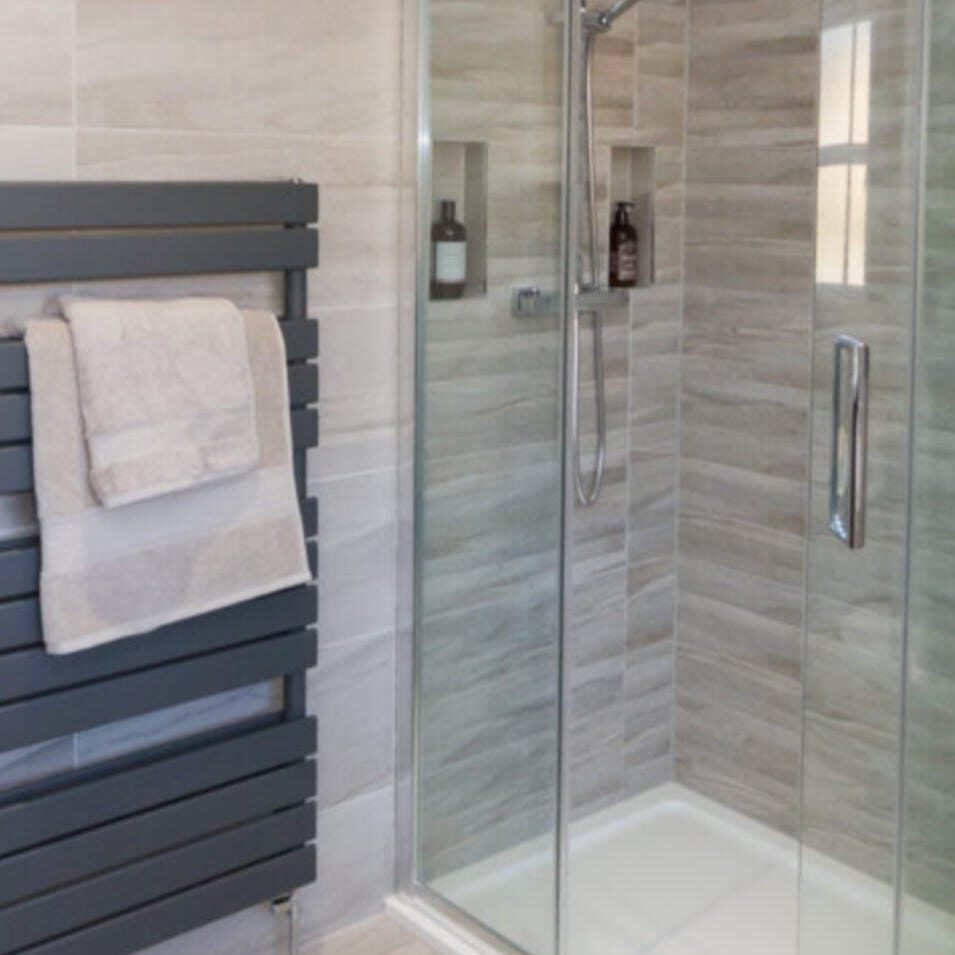 A modern bathroom featuring a glass-enclosed shower with a wall-mounted rainfall showerhead. The shower has two built-in shelves with toiletries. Next to it is a dark gray heated towel rack with folded white towels. The walls are tiled in beige.