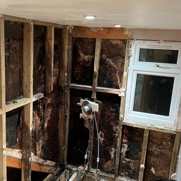 A room with exposed wooden studs and severely damaged, burnt walls. There is a partially intact window and two ceiling lights. The space appears to be under renovation or repair.